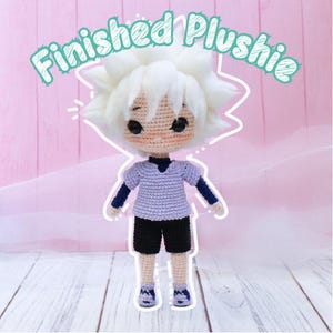 Op de afbeelding: Een gehaakt pluche speelgoed van een personage met wit haar, een paars shirt, een zwarte broek en blauwe schoenen. Het speelgoed staat op een wit houten oppervlak. De tekst "Finished Plushie" is geschreven in een witte omtrek op een roze achtergrond.