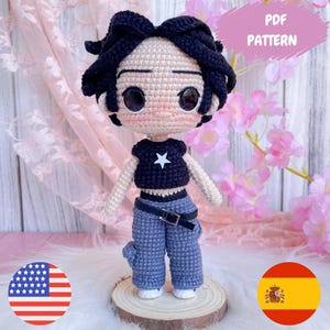 Puede incluir: Muñeca de ganchillo con pelo negro y ojos grandes, vestida con un top negro con una estrella blanca, pantalones azules y un cinturón negro. La muñeca está sobre una base de madera. Incluye el texto "PDF PATTERN".
