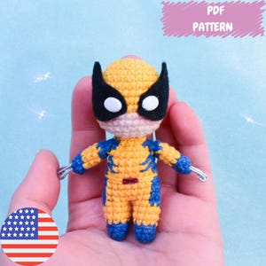 Puede incluir: Juguete amigurumi de crochet de un personaje de superhéroe con ropa amarilla y azul, máscara negra y ojos blancos. El juguete está siendo sostenido en una mano. La imagen incluye el texto "PDF PATTERN".