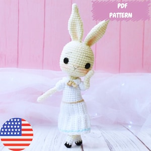 Puede incluir: Patrón de crochet para una muñeca de conejo blanco que lleva un vestido blanco con ribetes azules. El patrón está disponible para descargar en PDF. La muñeca está sentada sobre una superficie blanca con un fondo rosa.