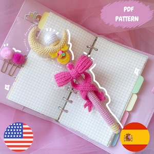 May include: A handmade crochet pen in pink and yellow, resembling a magical wand, rests on an open notebook. The pen features a pink bow and a yellow crescent moon design. The text "PDF PATTERN" is visible in the background.