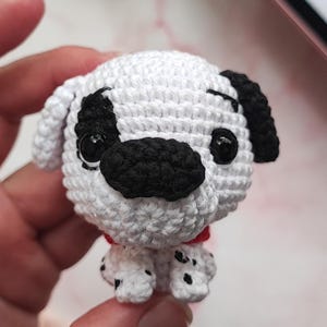 Puede incluir: Un peque&ntilde;o juguete de crochet de un perro d&aacute;lmata con manchas negras y un collar rojo. El juguete tiene ojos negros y una nariz negra.