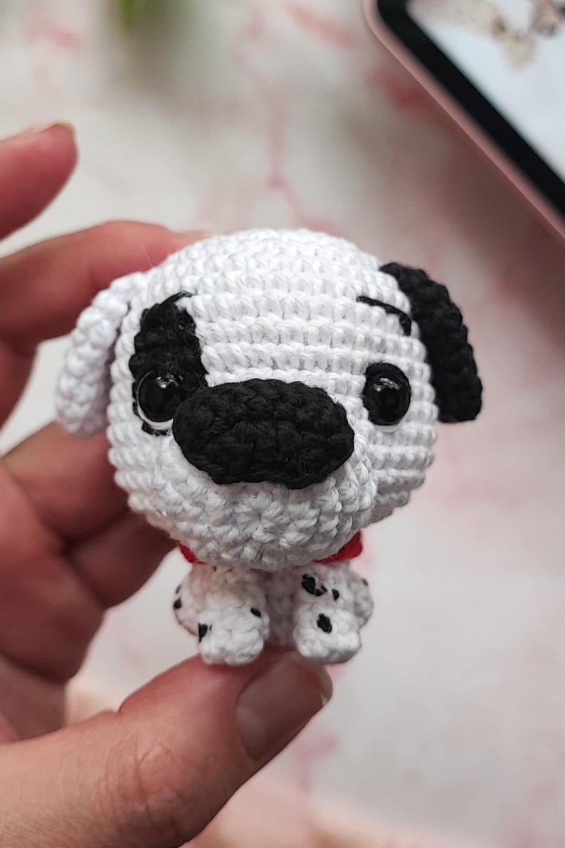 Puede incluir: Un peque&ntilde;o juguete de crochet de un perro d&aacute;lmata con manchas negras y un collar rojo. El juguete tiene ojos negros y una nariz negra.