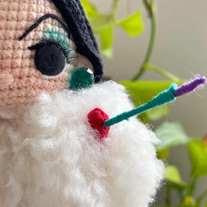 Puede incluir: Una mu&ntilde;eca de crochet con cabello negro, ojos verdes y barba blanca. La mu&ntilde;eca lleva un vestido negro y tiene una nariz roja.