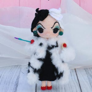 Puede incluir: Una mu&ntilde;eca de ganchillo de Cruella de Vil de la pel&iacute;cula de Disney 101 D&aacute;lmatas. La mu&ntilde;eca lleva un vestido negro y un abrigo de piel blanco. Tiene el pelo negro y blanco, sombra de ojos verde y zapatos rojos.