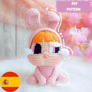 Puede incluir: Muñeca de ganchillo rosa con orejas de conejo y pelo naranja bajo la capucha. La muñeca tiene ojos negros y rosas, detalles blancos y está sentada. La imagen incluye el texto "PDF PATTERN".