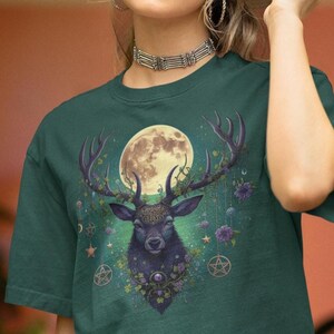 May include: A dark green t-shirt with a graphic print of a black stag with antlers, surrounded by flowers, vines, and a full moon. The stag has a jeweled necklace and a crown of leaves.