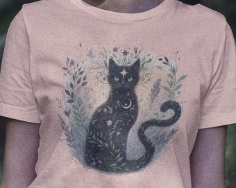 May include: A woman wearing a pink t-shirt with a black cat illustration. The cat is surrounded by foliage and has a crescent moon and stars on its body. The woman is standing in a forest setting.