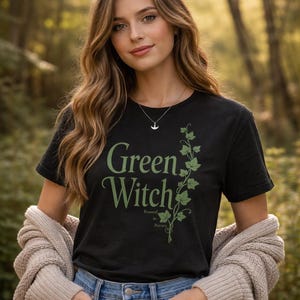 Peut inclure: T-shirt noir avec l'inscription "Green Witch" en vert et un motif de vigne verte. Le t-shirt porte &eacute;galement les mots "Rooted in Nature" en plus petit. La personne porte un jean bleu et un cardigan beige.