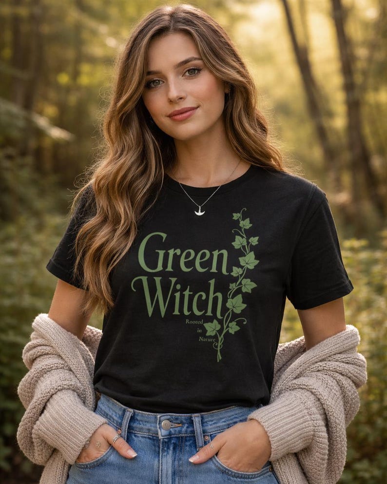 Peut inclure: T-shirt noir avec l'inscription "Green Witch" en vert et un motif de vigne verte. Le t-shirt porte &eacute;galement les mots "Rooted in Nature" en plus petit. La personne porte un jean bleu et un cardigan beige.