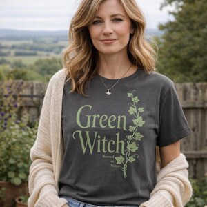Peut inclure: Un t-shirt gris fonc&eacute; avec l'inscription "Green Witch" en vert clair, accompagn&eacute; d'un motif de vigne. L'expression "Rooted in Nature" est en dessous. La personne porte un cardigan cr&egrave;me et un jean bleu.
