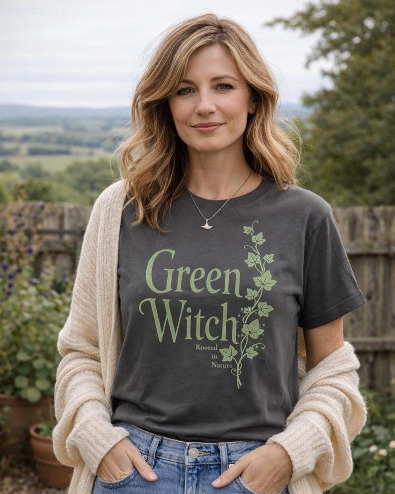 Peut inclure: Un t-shirt gris fonc&eacute; avec l'inscription "Green Witch" en vert clair, accompagn&eacute; d'un motif de vigne. L'expression "Rooted in Nature" est en dessous. La personne porte un cardigan cr&egrave;me et un jean bleu.