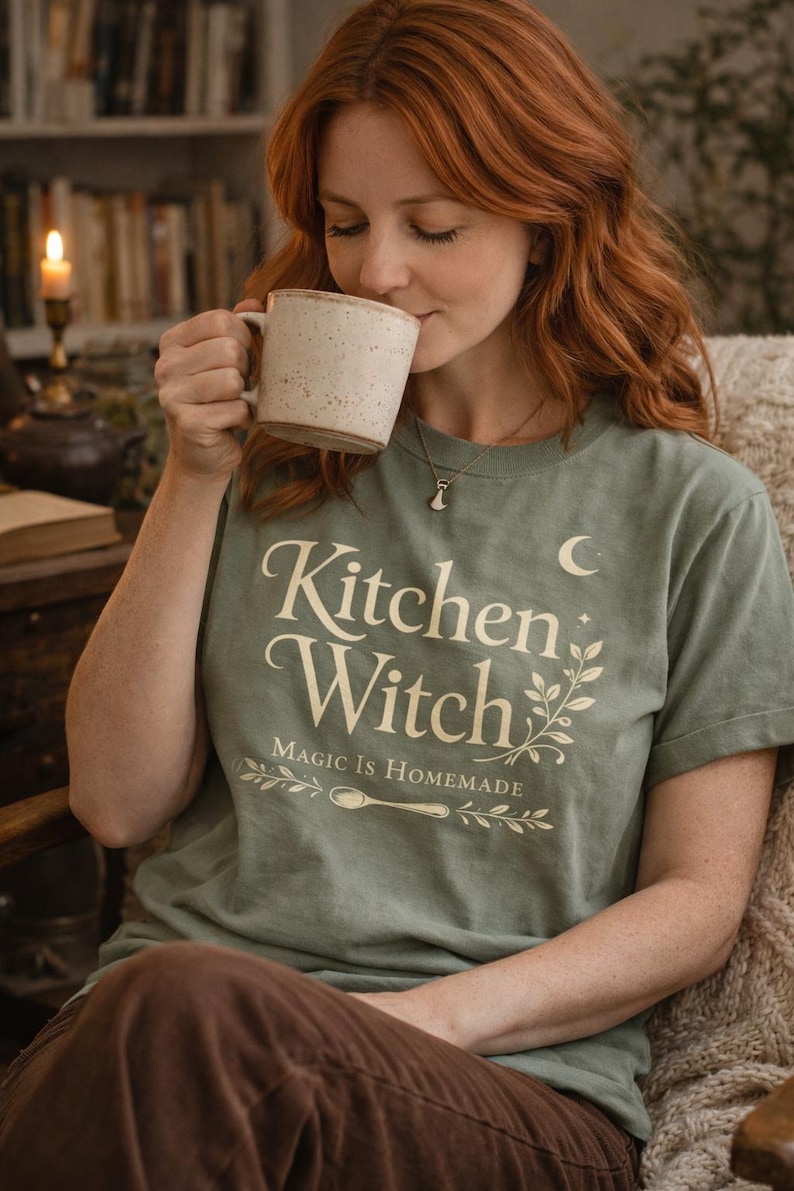 May include: Sage green t-shirt with the text "Kitchen Witch" in a decorative font, with the phrase "Magic is Homemade" below. The shirt is worn by a person holding a speckled ceramic mug.