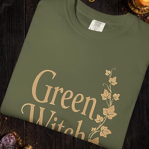 Peut inclure: T-shirt vert olive avec l'inscription "Green Witch" en lettres dor&eacute;es, accompagn&eacute;e d'un motif de feuilles et de vigne dor&eacute;es. Le mot "Hemp" est au-dessus en lettres dor&eacute;es.