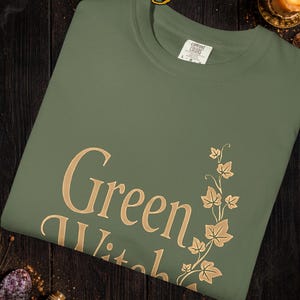 Peut inclure: T-shirt vert sauge avec l'inscription "Green Witch" en lettres dor&eacute;es, accompagn&eacute;e d'un motif de feuilles. Le t-shirt est en mati&egrave;re Comfort Colors. Le texte "Colors and graphics may vary slightly due to computer screens and printing differences" est &eacute;galement visible.