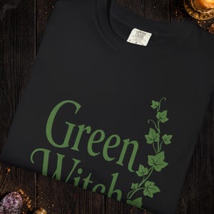 Peut inclure: Sweat-shirt noir avec l'inscription "Green Witch" en vert, accompagn&eacute;e d'un motif de lierre vert. Le mot "Black" est &eacute;crit en or en haut. Le texte "Colors and graphics may vary slightly" est en bas.