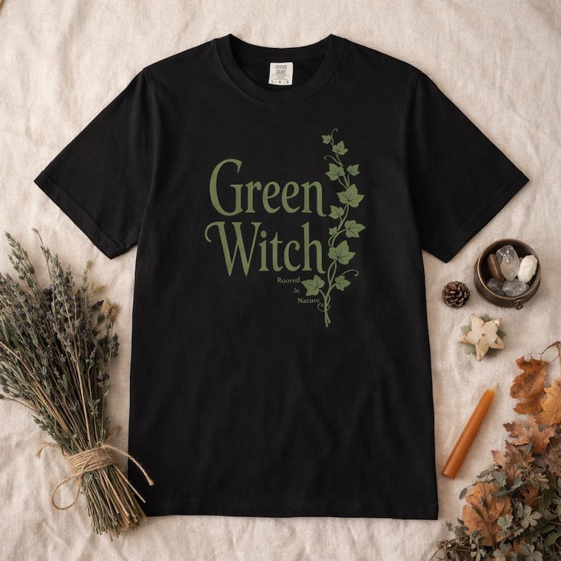 Peut inclure: T-shirt noir avec l'inscription "Green Witch" en vert, accompagn&eacute;e d'un motif de vigne verte. L'expression "Rooted in Nature" est en dessous. Le t-shirt est pr&eacute;sent&eacute; sur une surface neutre, avec des herbes s&eacute;ch&eacute;es et des objets d&eacute;coratifs.