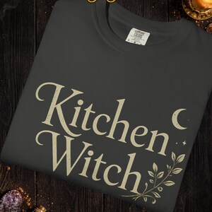 May include: Dark gray t-shirt with the words "Kitchen Witch" in a vintage font. The design includes a crescent moon, stars, and botanical accents. The shirt is a Comfort Colors brand.