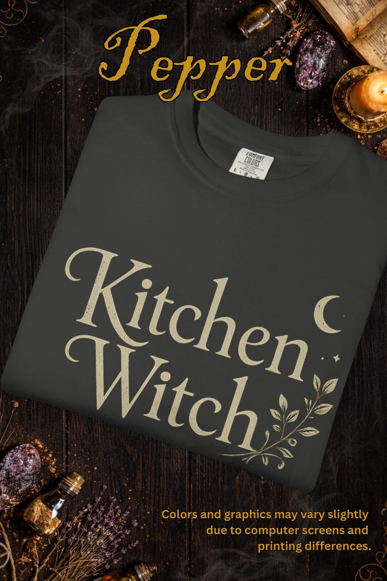 May include: Dark gray t-shirt with the words "Kitchen Witch" in a vintage font. The design includes a crescent moon, stars, and botanical accents. The shirt is a Comfort Colors brand.