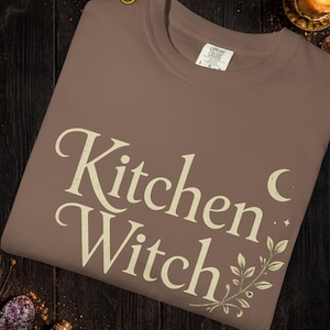 May include: A brown t-shirt with the words "Kitchen Witch" in a cream-colored, vintage-style font. The word "Espresso" is above the shirt. The shirt has a crew neck and short sleeves. The design includes a crescent moon and botanical elements.