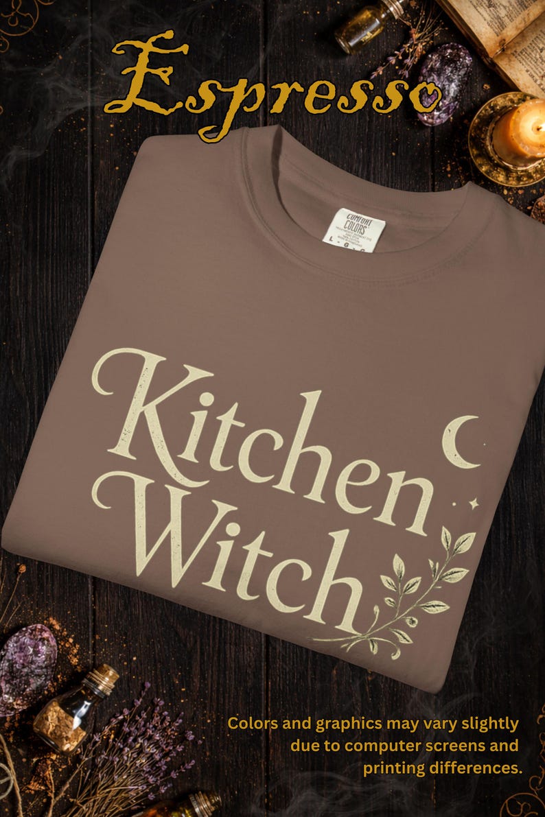 May include: A brown t-shirt with the words "Kitchen Witch" in a cream-colored, vintage-style font. The word "Espresso" is above the shirt. The shirt has a crew neck and short sleeves. The design includes a crescent moon and botanical elements.
