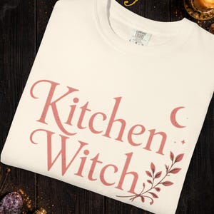 May include: Ivory-colored t-shirt with the words "Kitchen Witch" in a pink script font. The design includes a crescent moon, stars, and botanical accents. The shirt is made by Comfort Colors.