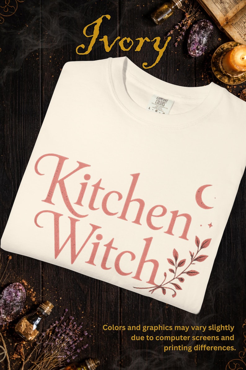 May include: Ivory-colored t-shirt with the words "Kitchen Witch" in a pink script font. The design includes a crescent moon, stars, and botanical accents. The shirt is made by Comfort Colors.
