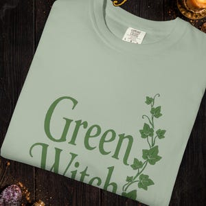 Peut inclure: T-shirt vert clair avec l'inscription "Green Witch" en vert fonc&eacute;, accompagn&eacute;e d'un motif de lierre vert. Le t-shirt est de la marque Comfort Colors. Le mot "Bay" est en haut. Le texte en bas indique que les couleurs peuvent varier.