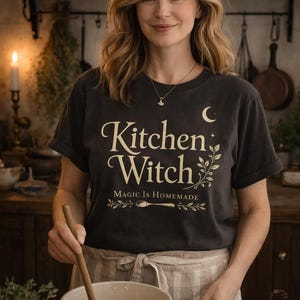 May include: Dark gray t-shirt with the words "Kitchen Witch" in a decorative font, along with the phrase "Magic is Homemade." The shirt features a crescent moon and botanical elements. A woman is wearing the shirt while cooking.