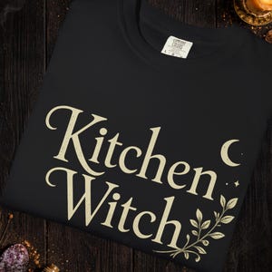 May include: Black crew neck sweatshirt with the text "Kitchen Witch" in a cream-colored, stylized font. The design includes a crescent moon and botanical accents. The word "Black" is at the top. The bottom of the image has text about color variations.