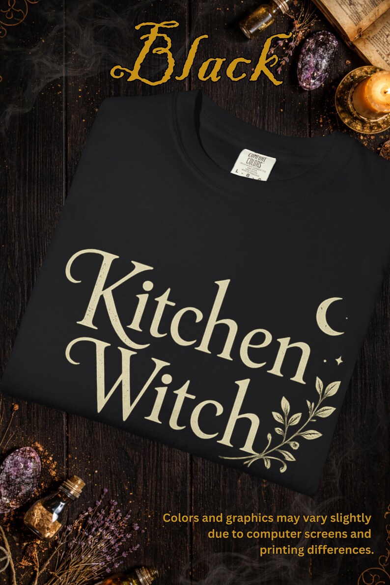 May include: Black crew neck sweatshirt with the text "Kitchen Witch" in a cream-colored, stylized font. The design includes a crescent moon and botanical accents. The word "Black" is at the top. The bottom of the image has text about color variations.