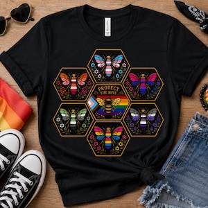 Op de afbeelding: Een zwart t-shirt met een afbeelding van zeven zeshoekige cellen in een cluster. Elke cel bevat een bij met vleugels in de kleuren van diverse pride-vlaggen. De middelste zeshoek toont een bij en de tekst Protect The Hive.