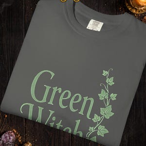 Peut inclure: Un t-shirt gris fonc&eacute; avec les mots "Green Witch" en vert clair, accompagn&eacute; d'un motif de lierre vert. Le mot "Pepper" est en haut. Le t-shirt est pli&eacute; sur une surface en bois sombre.