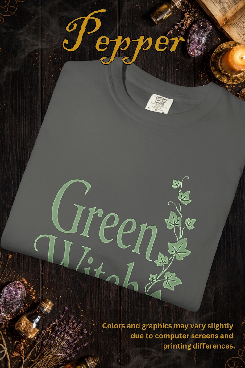 Peut inclure: Un t-shirt gris fonc&eacute; avec les mots "Green Witch" en vert clair, accompagn&eacute; d'un motif de lierre vert. Le mot "Pepper" est en haut. Le t-shirt est pli&eacute; sur une surface en bois sombre.