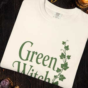 Peut inclure: T-shirt ivoire avec l'inscription "Green Witch" en vert et un motif de lierre vert. Le t-shirt est en mati&egrave;re douce. Le texte "Colors and graphics may vary slightly due to computer screens and printing differences" est &eacute;galement visible.