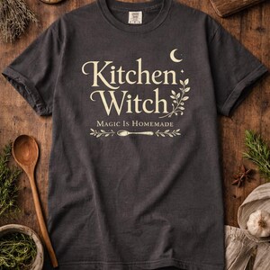 May include: Dark gray t-shirt with the words "Kitchen Witch" in a decorative font, along with the phrase "Magic is Homemade." The design includes a crescent moon, sprigs, and a spoon. The shirt is displayed on a wooden surface with herbs and spices.
