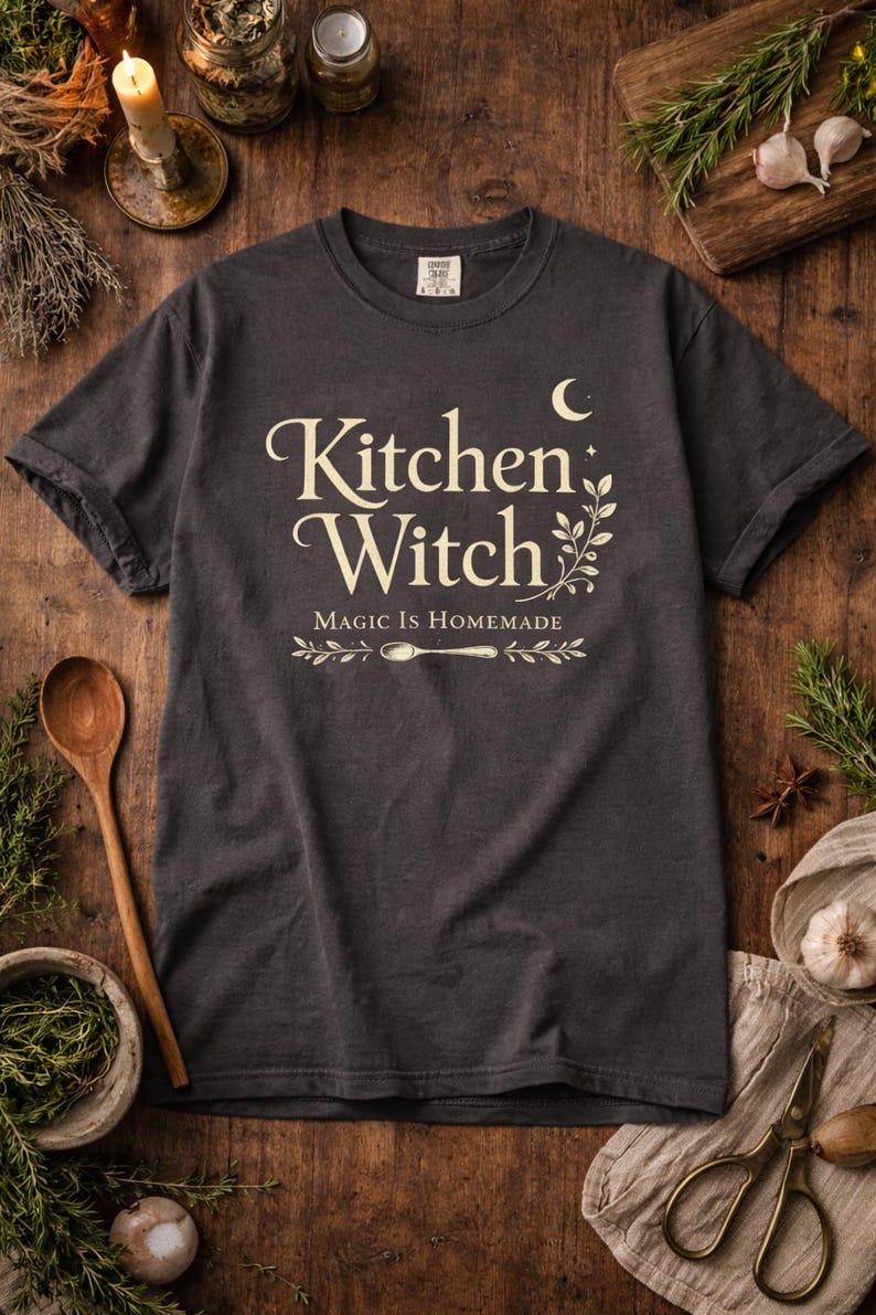 May include: Dark gray t-shirt with the words "Kitchen Witch" in a decorative font, along with the phrase "Magic is Homemade." The design includes a crescent moon, sprigs, and a spoon. The shirt is displayed on a wooden surface with herbs and spices.