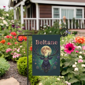 Puede incluir: Una bandera de jard&iacute;n verde con una silueta de ciervo negro, una luna llena y la palabra "Beltane" en letras doradas. El ciervo est&aacute; rodeado de flores y enredaderas.