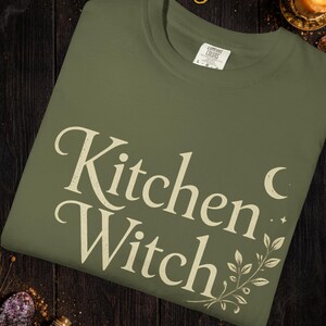 May include: Olive green t-shirt with the words "Kitchen Witch" in a vintage font, accented with a crescent moon and botanical illustration. The word "Hemp" is at the top. The shirt is made of Comfort Colors material.