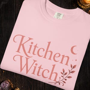 May include: Light pink t-shirt with the text "Kitchen Witch" in a stylized font. The design includes a crescent moon, stars, and botanical elements. The shirt is from Comfort Colors.