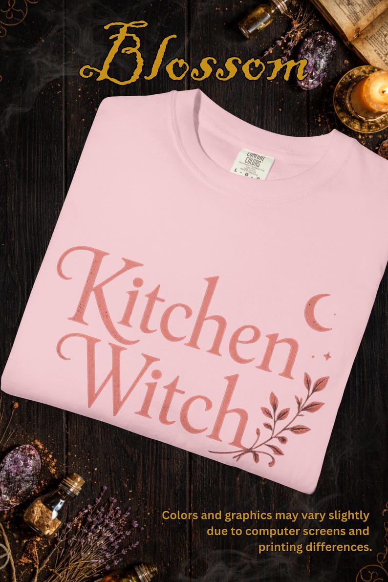 May include: Light pink t-shirt with the text "Kitchen Witch" in a stylized font. The design includes a crescent moon, stars, and botanical elements. The shirt is from Comfort Colors.
