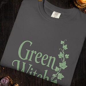 Peut inclure: T-shirt graphite avec l'inscription "Green Witch" en vert clair, accompagn&eacute;e d'un motif botanique. Le t-shirt est en mati&egrave;re douce et pr&eacute;sente un col rond. Le texte "Colors and graphics may vary slightly due to computer screens and printing differences" est &eacute;galement visible.