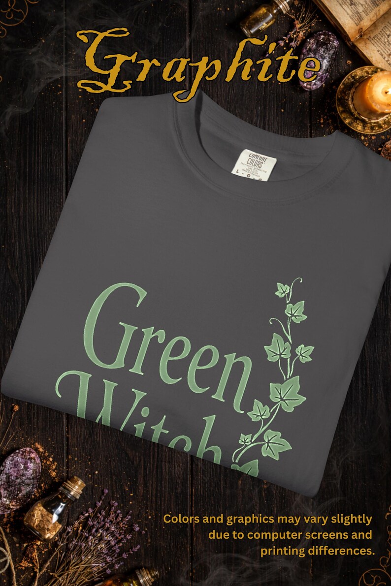 Peut inclure: T-shirt graphite avec l'inscription "Green Witch" en vert clair, accompagn&eacute;e d'un motif botanique. Le t-shirt est en mati&egrave;re douce et pr&eacute;sente un col rond. Le texte "Colors and graphics may vary slightly due to computer screens and printing differences" est &eacute;galement visible.