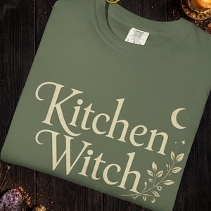 May include: Sage green t-shirt with the words "Kitchen Witch" in a vintage font. The design includes a crescent moon, stars, and botanical accents. The shirt is a Comfort Colors brand.