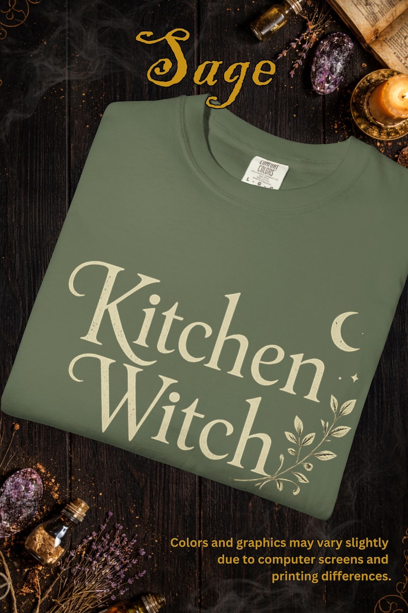 May include: Sage green t-shirt with the words "Kitchen Witch" in a vintage font. The design includes a crescent moon, stars, and botanical accents. The shirt is a Comfort Colors brand.