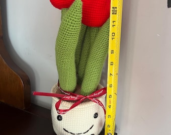 May include: A crocheted flower pot with a smiling face and red tulips. The pot has a red ribbon tied around it with the text "Just for you" on it.