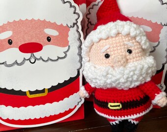May include: A crocheted Santa Claus doll with a white beard and a red hat. The doll is wearing a red and white suit with a black belt. The doll is sitting on a table in front of a red and white gift bag.