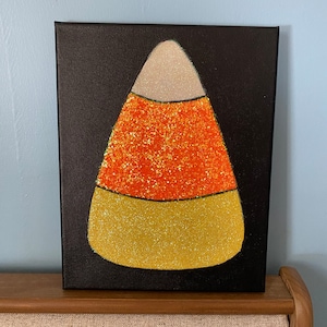 May include: A black canvas painting of a candy corn with orange, yellow, and white glitter.