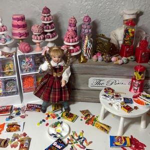 May include: Miniature candy shop display with a doll in a plaid dress. The display includes candy jars, cakes, lollipops, and packaged candies. A small white table holds more candy and treats. The sign on the display reads "The Petite Boutique."
