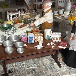 May include: A miniature kitchen scene featuring two dolls, one in a brown apron and one in a black and white uniform, standing at a wooden table. The table holds silver bowls, flour bags, egg cartons, a milk jug, measuring cups, and a small red book.